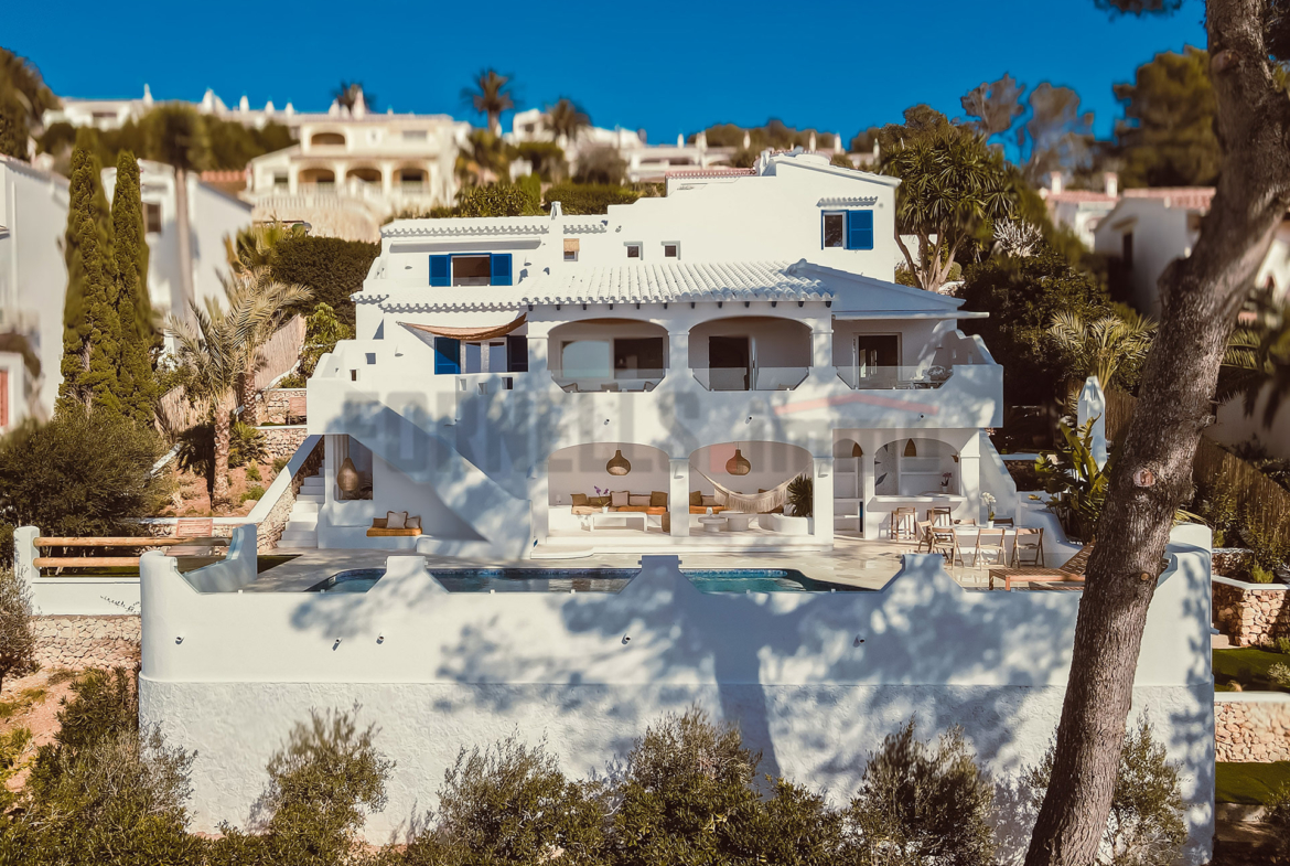 Fachada blanca de villa de lujo en Menorca con múltiples terrazas, piscina, vistas al mar y rodeada de vegetación mediterránea.