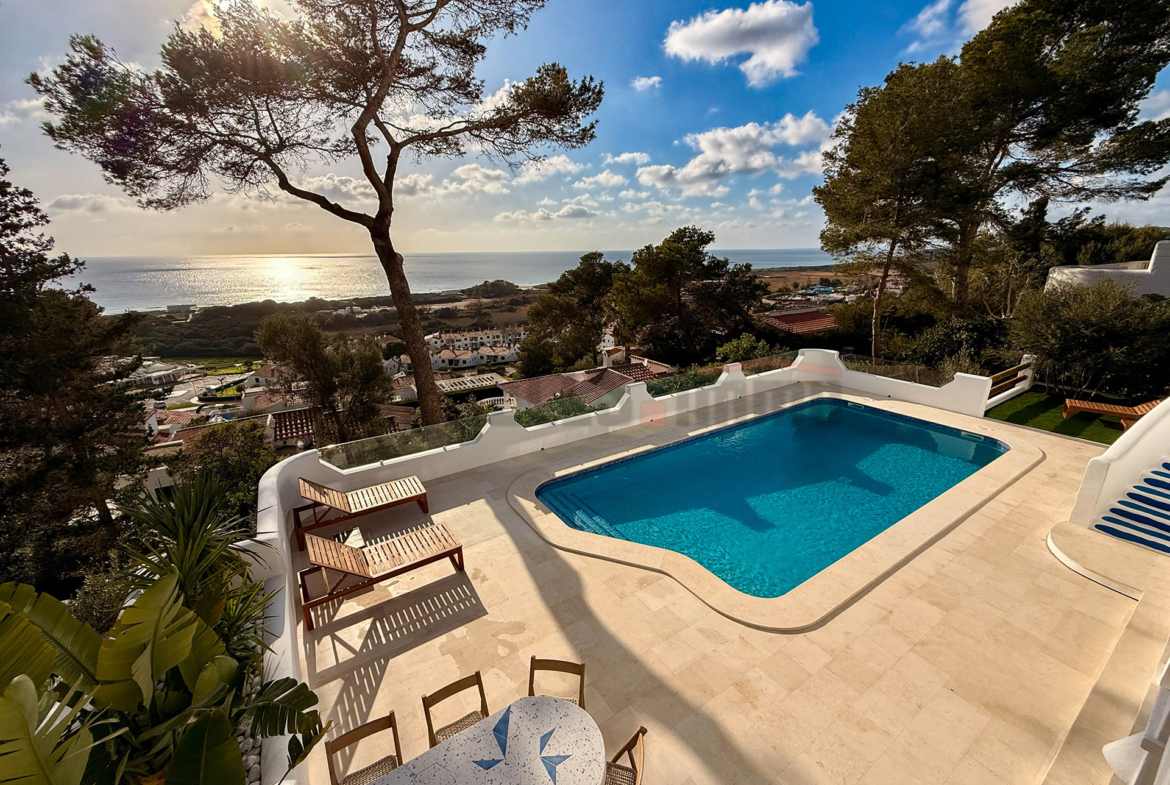 Piscina exterior de aguas azules en villa de lujo con solárium, rodeada de pinos y vistas al mar Mediterráneo en Menorca.