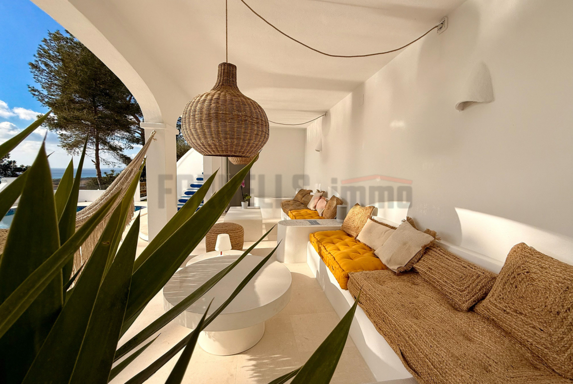 Terraza cubierta con sofás de obra, cojines amarillos y lámparas de mimbre, con vistas al exterior en villa de lujo en Menorca