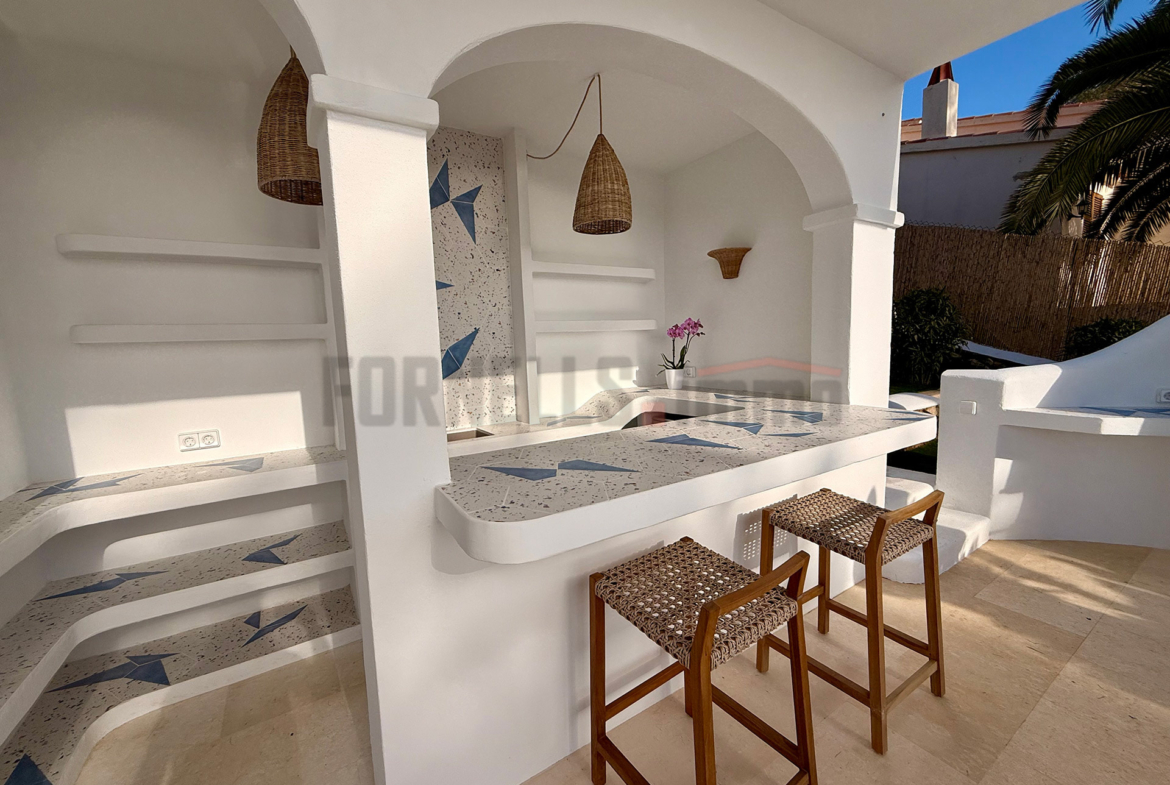 Barra de bar exterior con encimera de diseño, taburetes, estanterías integradas y lámparas de mimbre en la villa de lujo en Menorca.