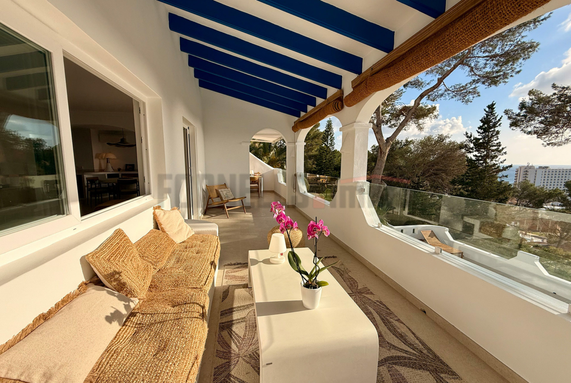 Terraza cubierta en primera planta con sofá, mesa de centro, vigas azules y vistas al exterior en villa de lujo en Menorca.
