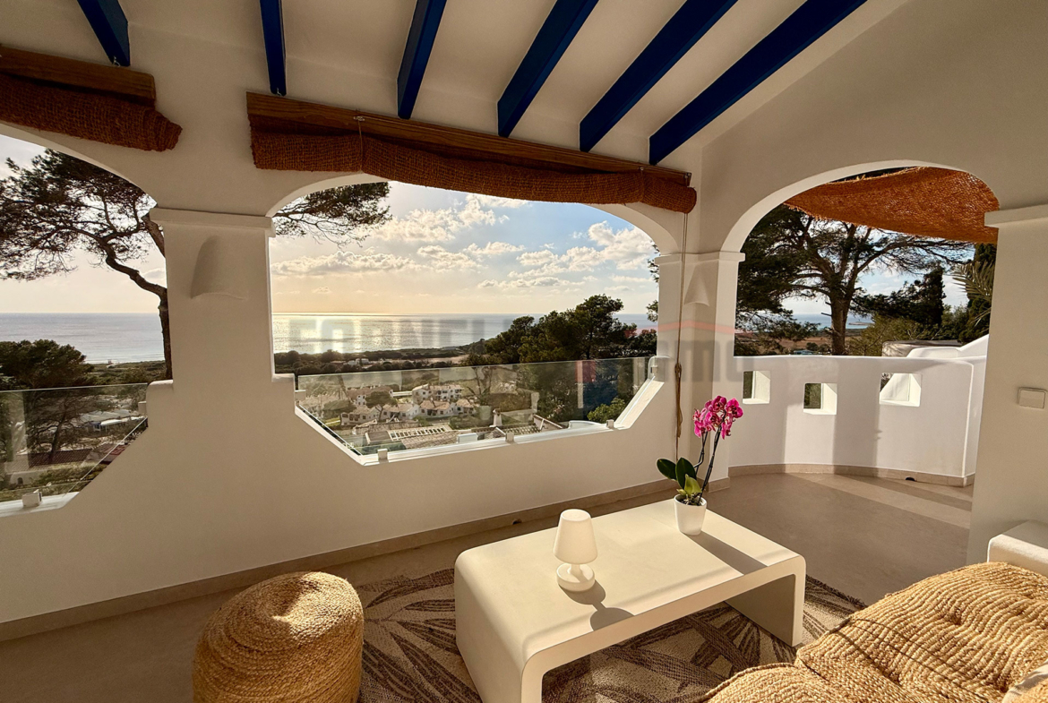Amplia terraza cubierta con área de estar, mesa de centro, puf, orquídea, y vistas panorámicas al mar y al paisaje en Menorca.