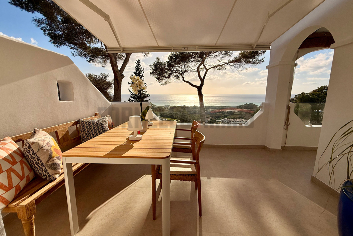 Mesa de comedor exterior de madera y metal, con banco y sillas, en terraza cubierta con vistas al mar en Menorca.