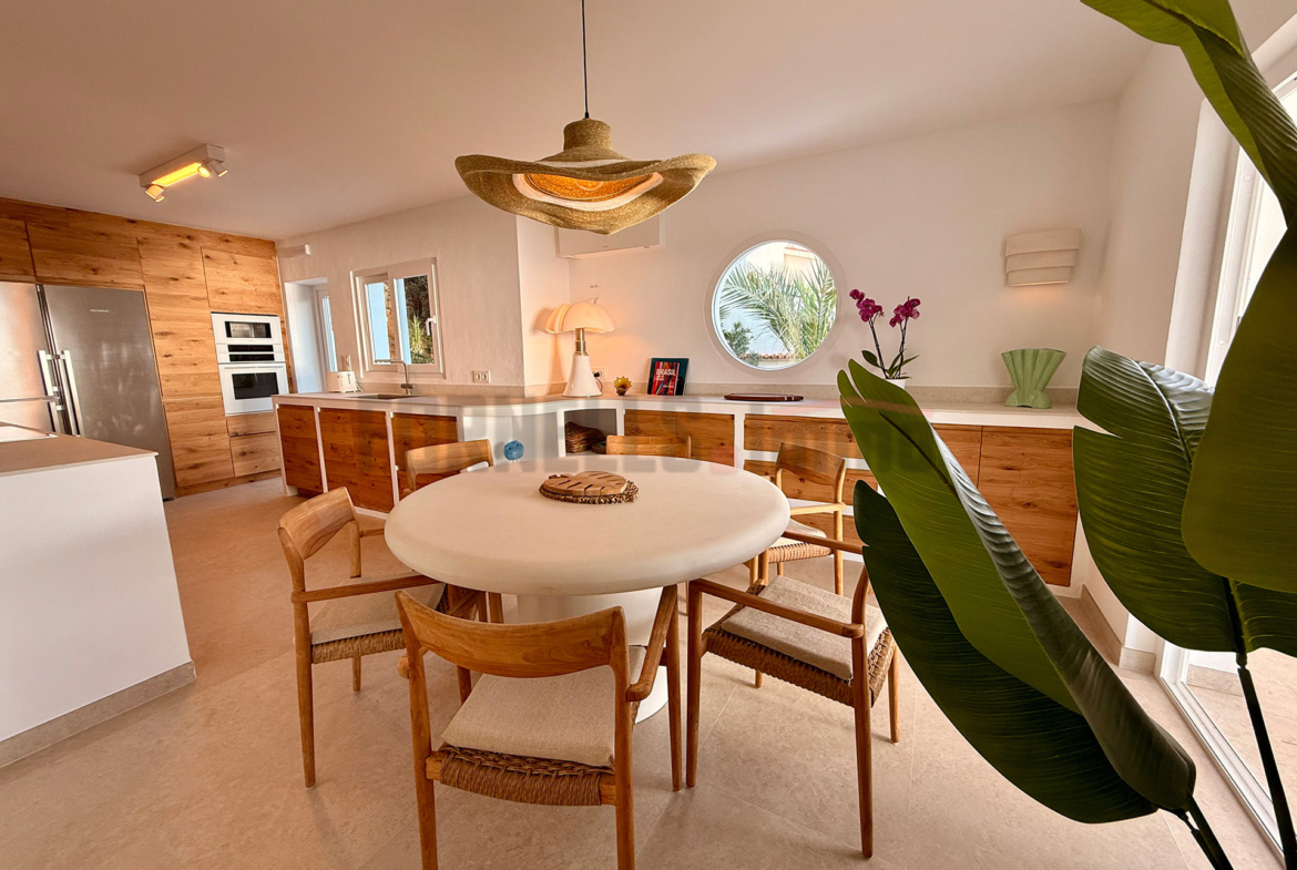 Amplio salón comedor con cocina integrada, mesa redonda, sillas de madera, lámpara de techo y decoración moderna en villa de lujo en Menorca.