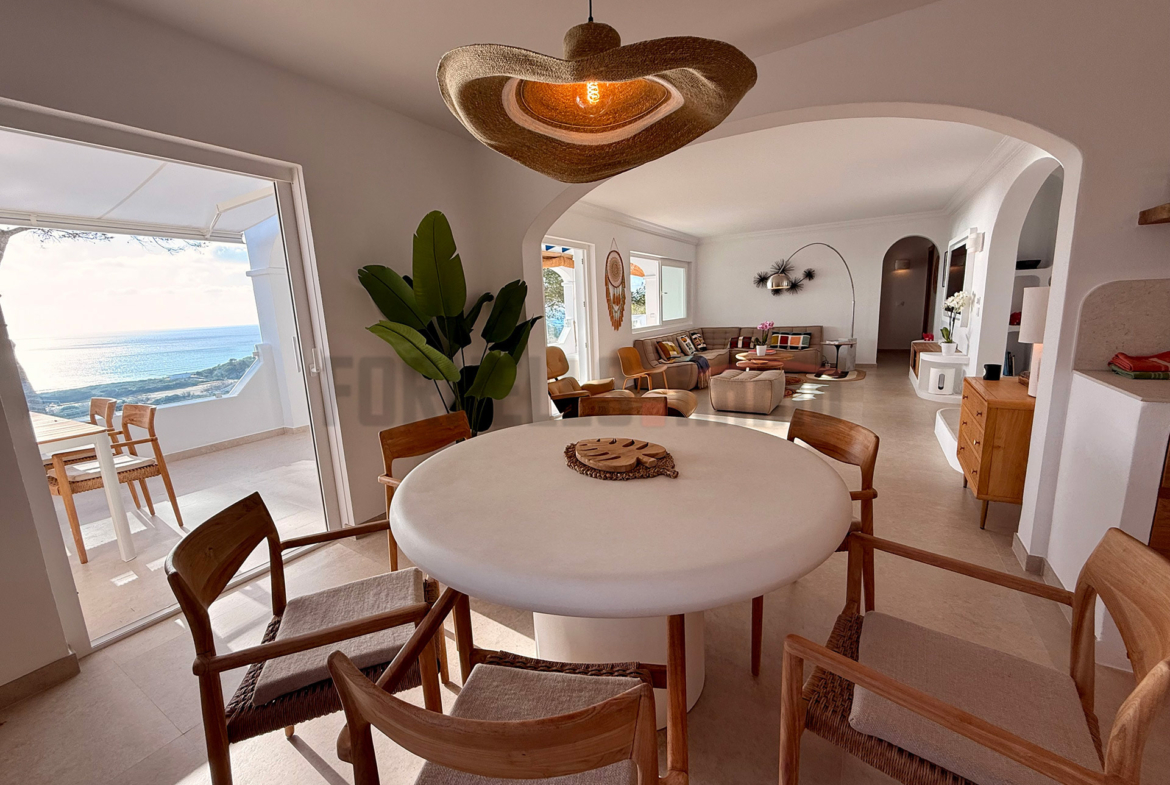 Amplio salón comedor de diseño con mesa redonda, sillas de madera, lámpara de techo y vistas al mar en villa de lujo en Menorca.