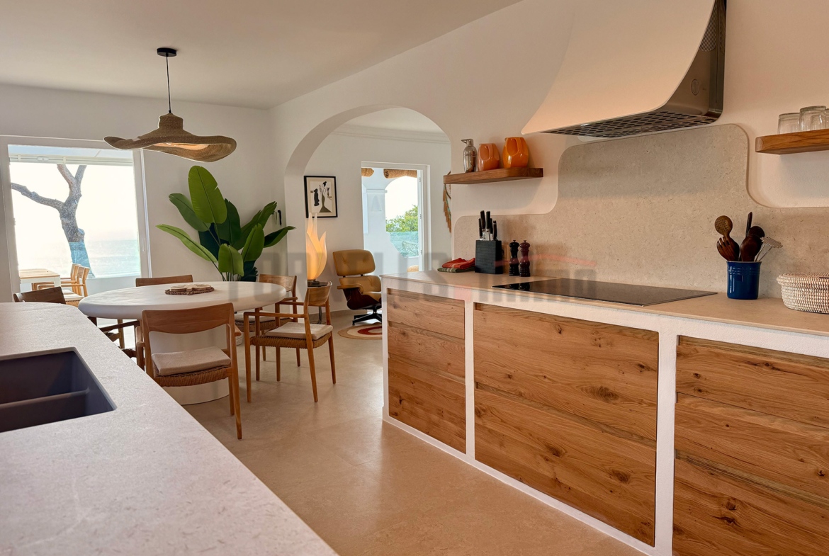 Cocina moderna con encimeras claras, armarios de madera, comedor integrado, y vistas al exterior en villa de lujo en Menorca.