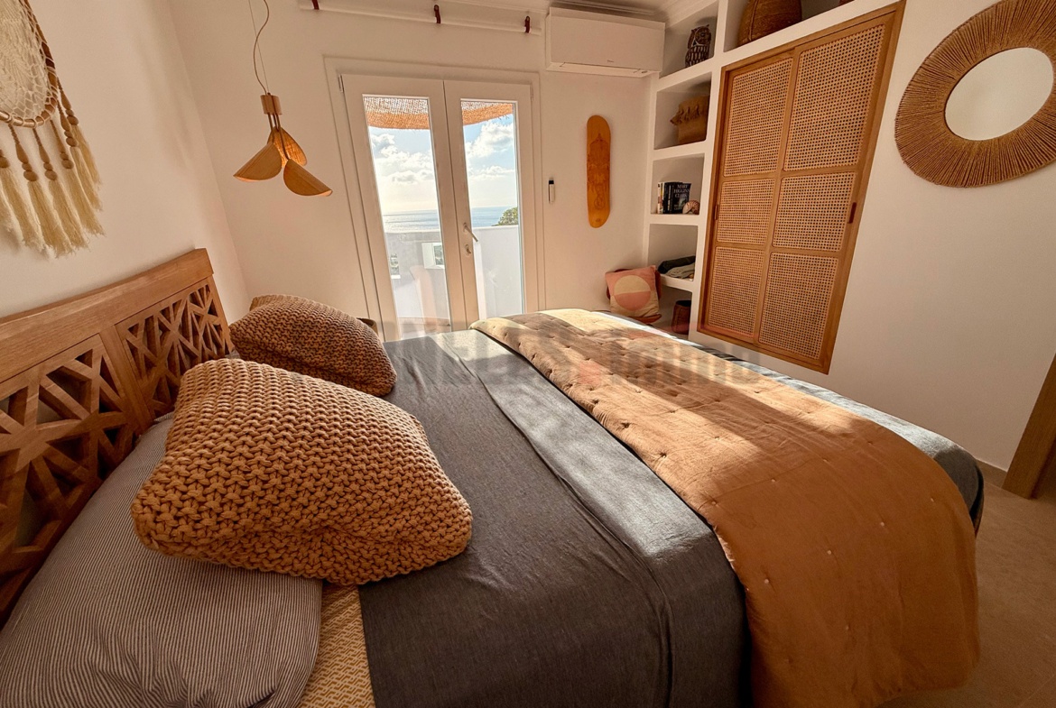 Dormitorio doble con cama de madera, textiles naturales, lavabo integrado, y acceso a balcón con vistas al mar en villa de lujo en Menorca.