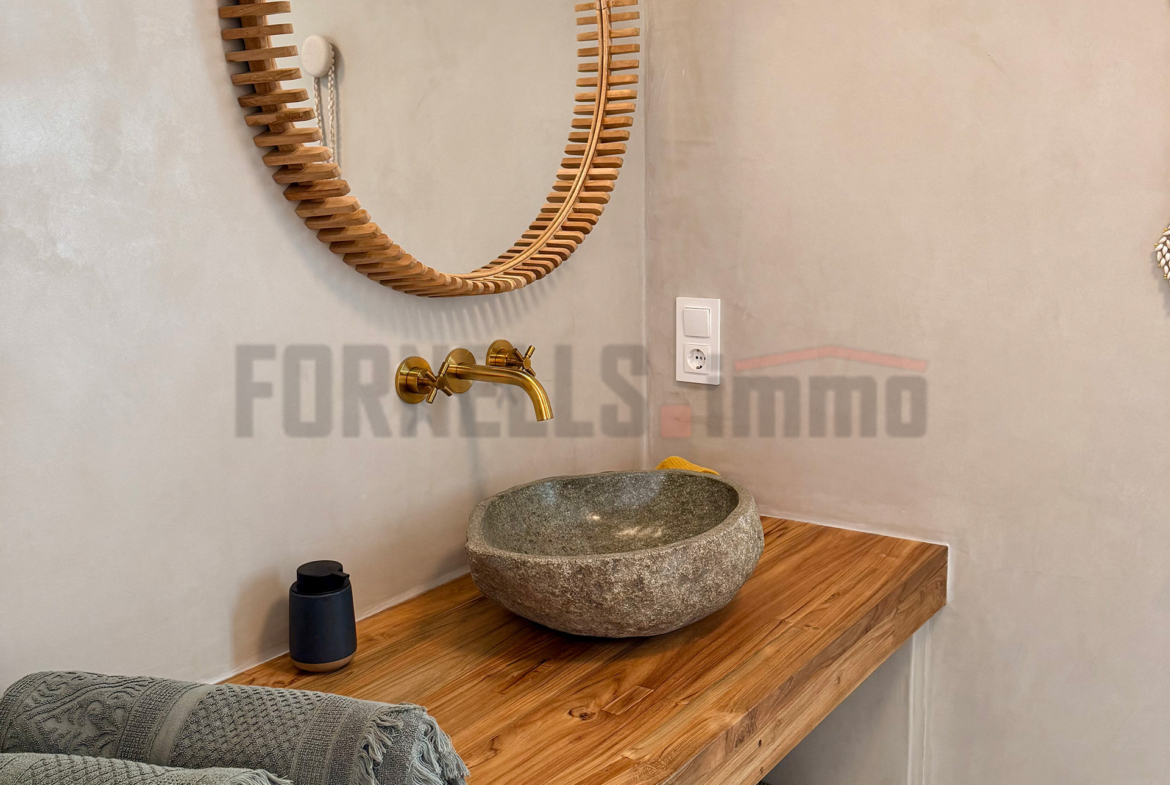 Detalle de lavabo de piedra, grifo dorado, espejo redondo de madera y toallas en baño de villa de lujo en Menorca.