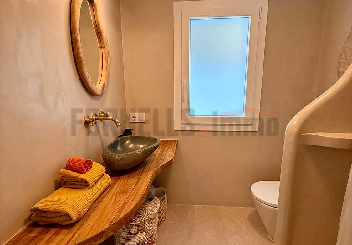 Lavabo de piedra natural sobre encimera de madera flotante, con espejo orgánico y toallas en baño de villa de lujo en Menorca.