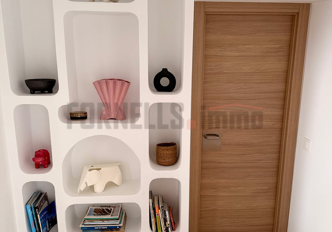 Estantería de obra blanca con nichos decorativos, objetos de arte y libros, junto a puerta de madera en pasillo de villa de lujo en Menorca.