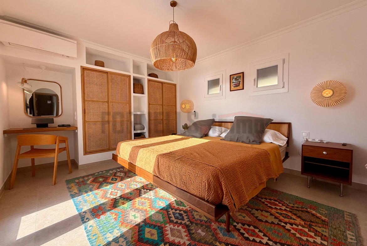 Amplia suite en planta superior con cama doble, armario de rejilla, escritorio, lavabo integrado, y decoración boho-chic en villa de lujo en Menorca.