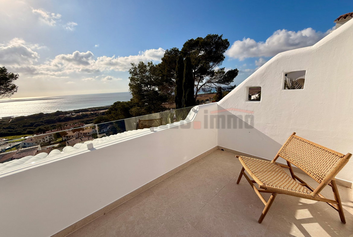 Terraza privada con sillón de mimbre, barandilla de cristal y vistas panorámicas al mar y al paisaje en villa de lujo en Menorca.