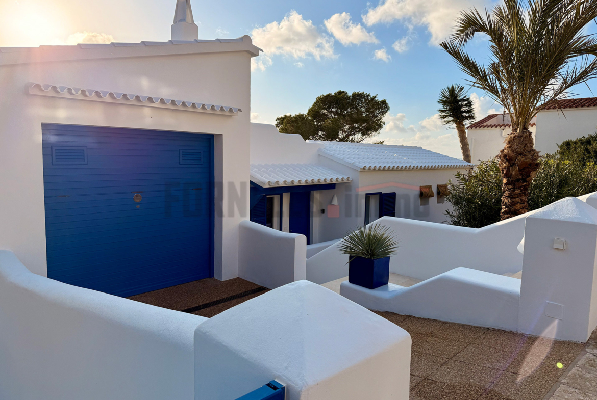 Acceso al garaje de villa de lujo con puerta azul vibrante, paredes blancas y vegetación mediterránea en Menorca.